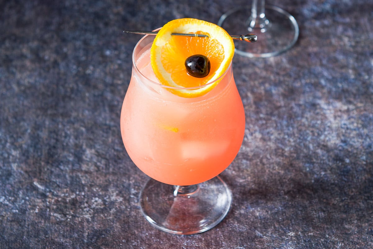A tulip glass with the cocktail with a shorter glass behind it and orange cup garnish with a cherry in each cup