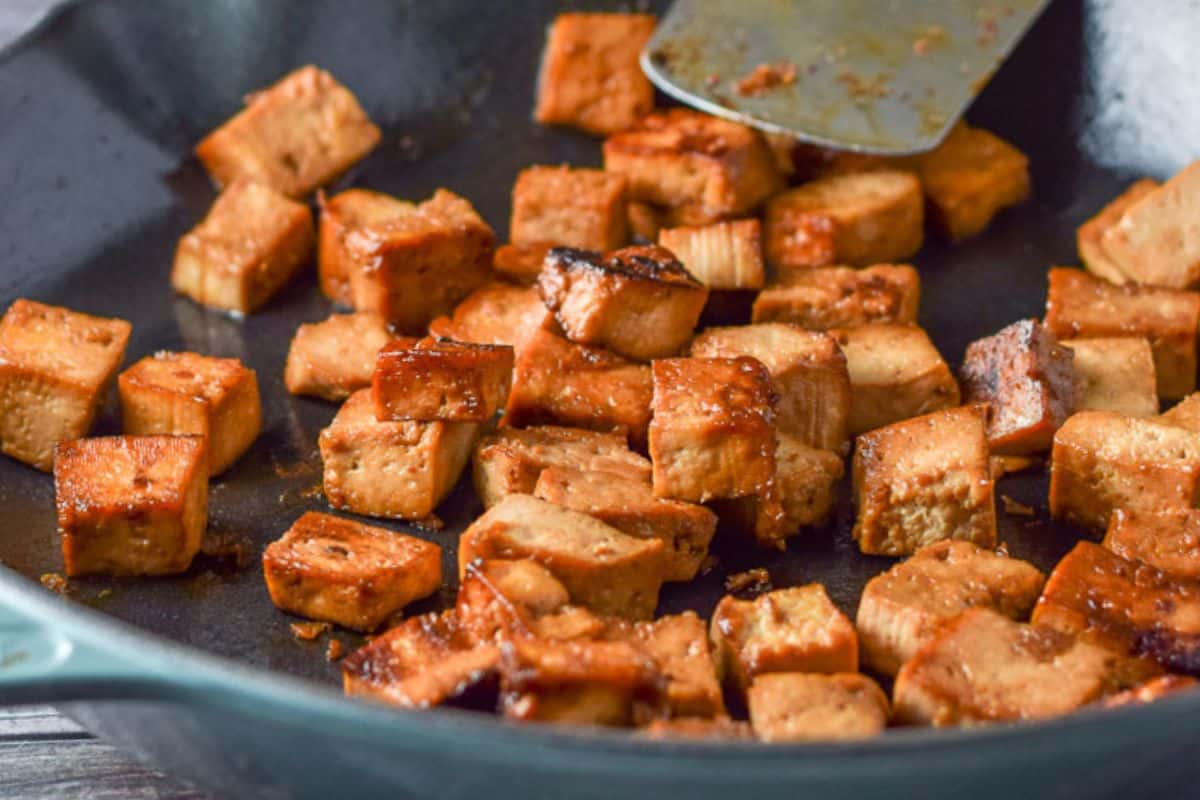 A blue cast iron pan with tofu that has been sautéed. There are some noodles and a salad in the background