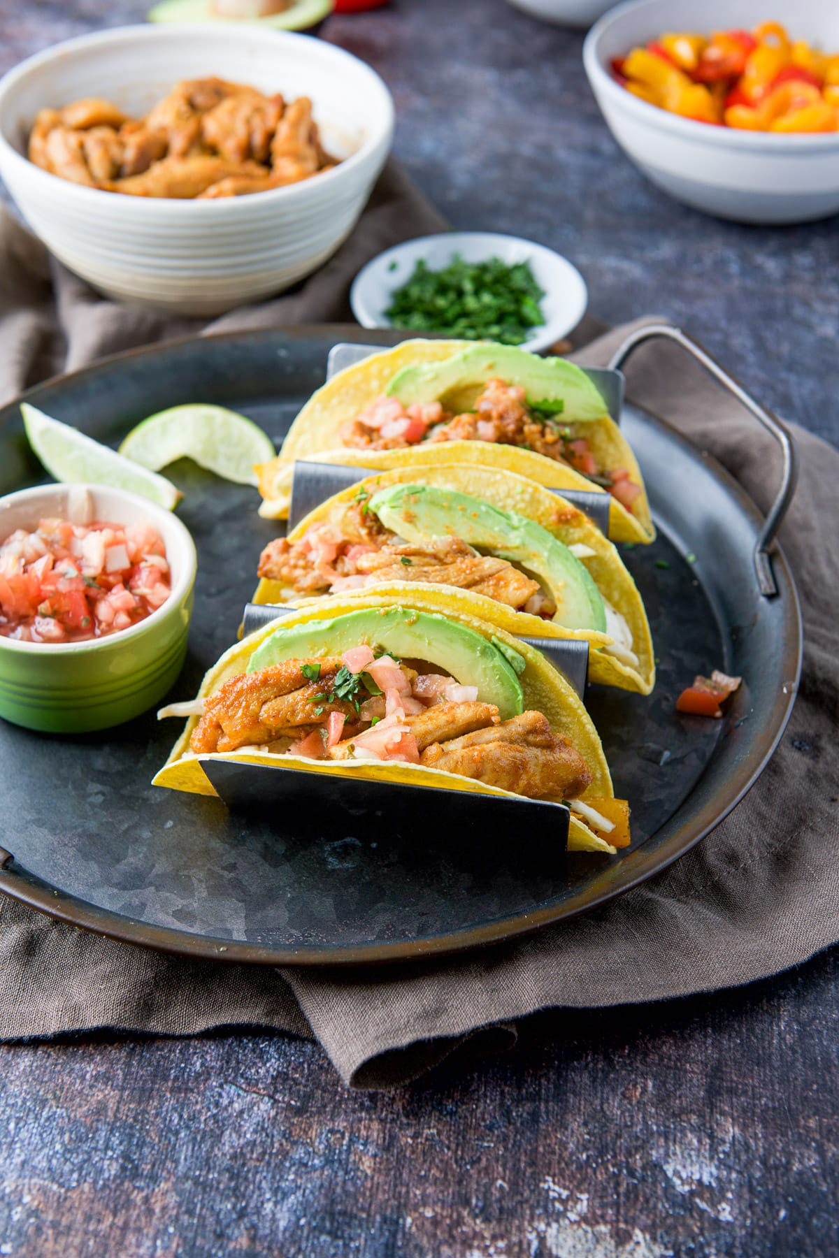 A metal plate with three tacos with chicken, avocado slices and pico de gallo, there are more ingredients in the back