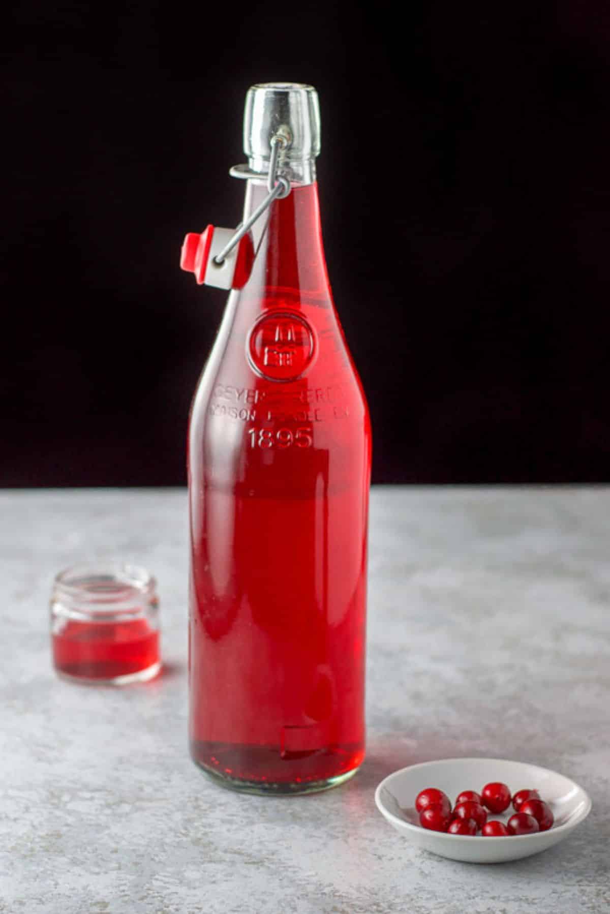 A glass bottle with cranberry vodka in it with more in a little jar and cranberries in a little white bowl