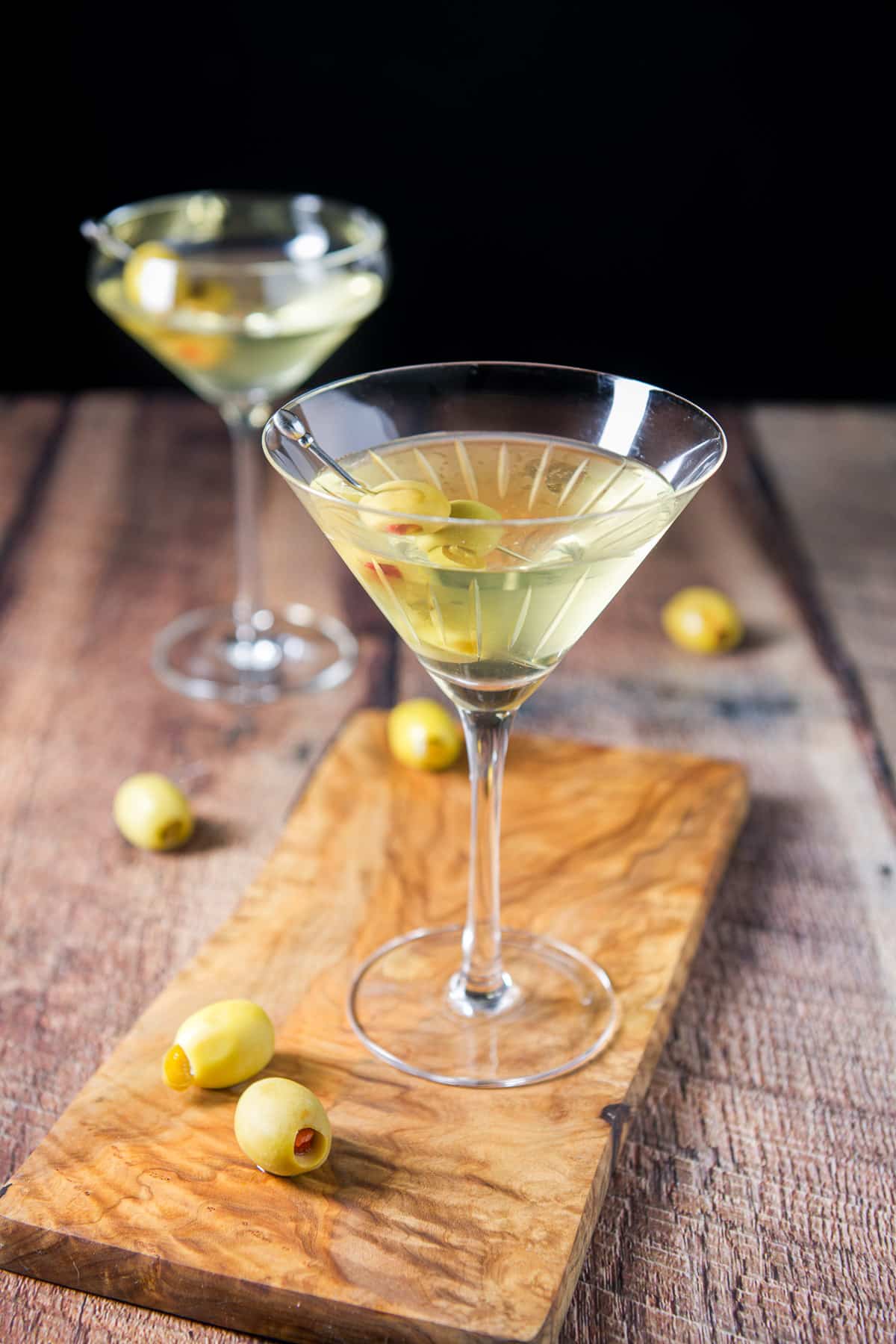 A beveled martini glass on a small board with another glass in the back, with olives as garnish and on the table