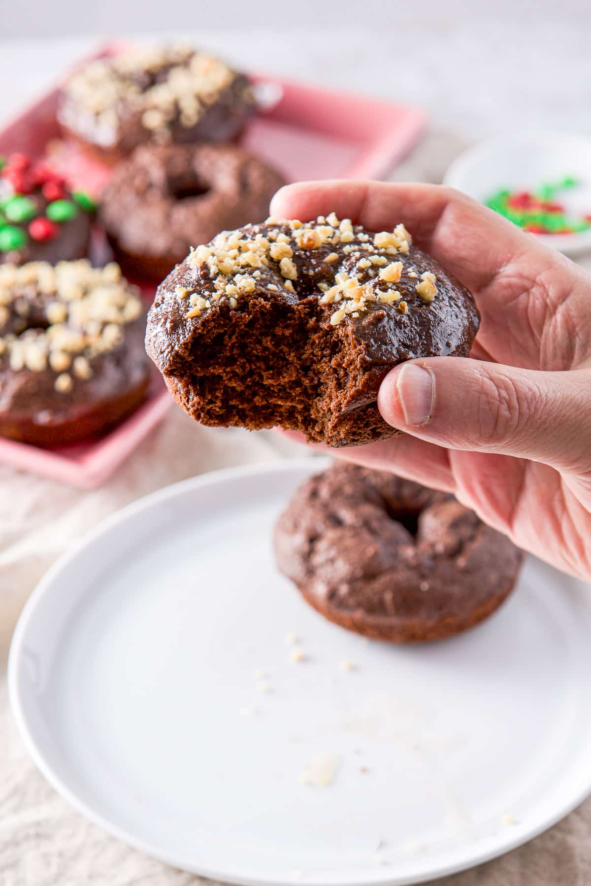A male hand holding a chocolate donut with walnuts on it. It's held over a white plate and there is a pink platter in the back
