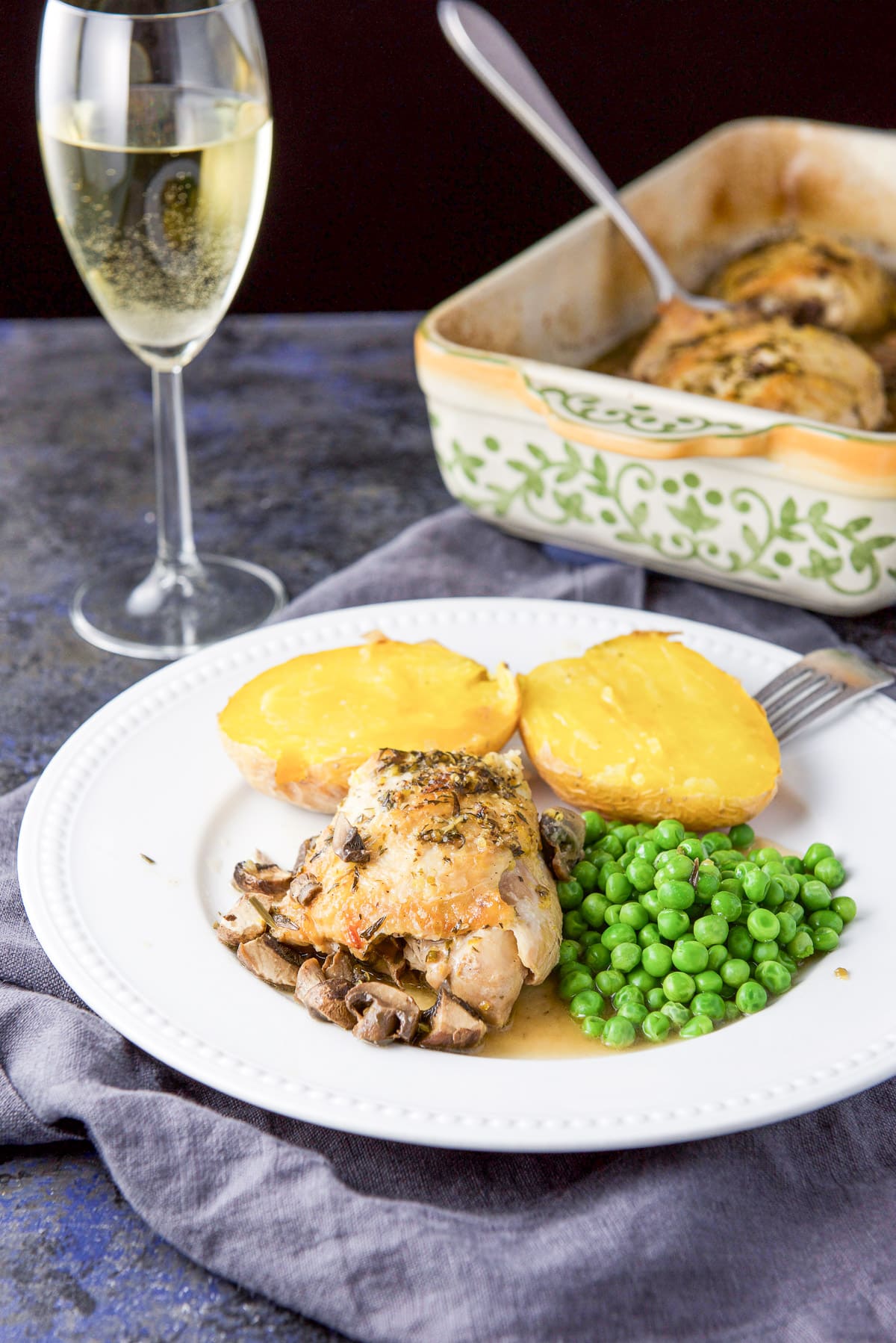 A beaded plate with a chicken thigh with sauce on it along with peas and a potato. There is a baking dish in the background
