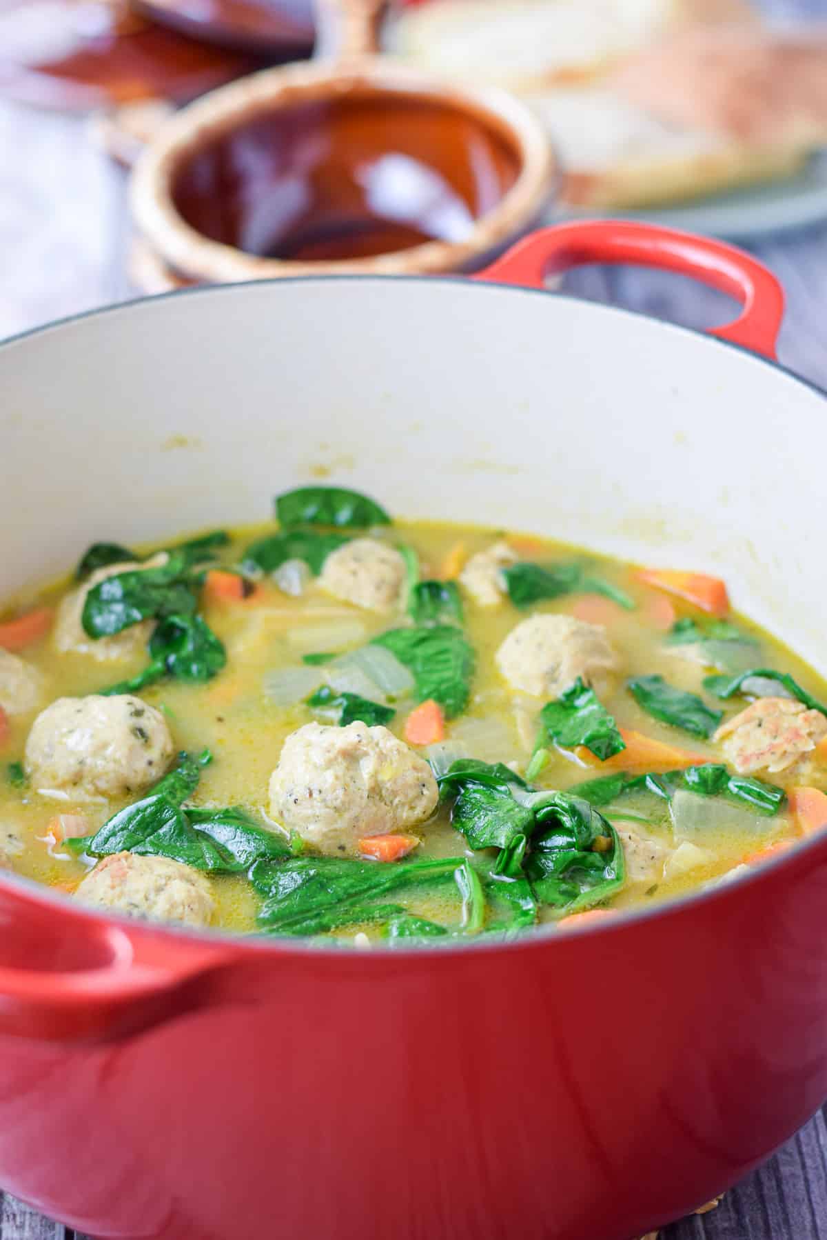 A red Dutch oven filled with a soup with meatballs and spinach in it
