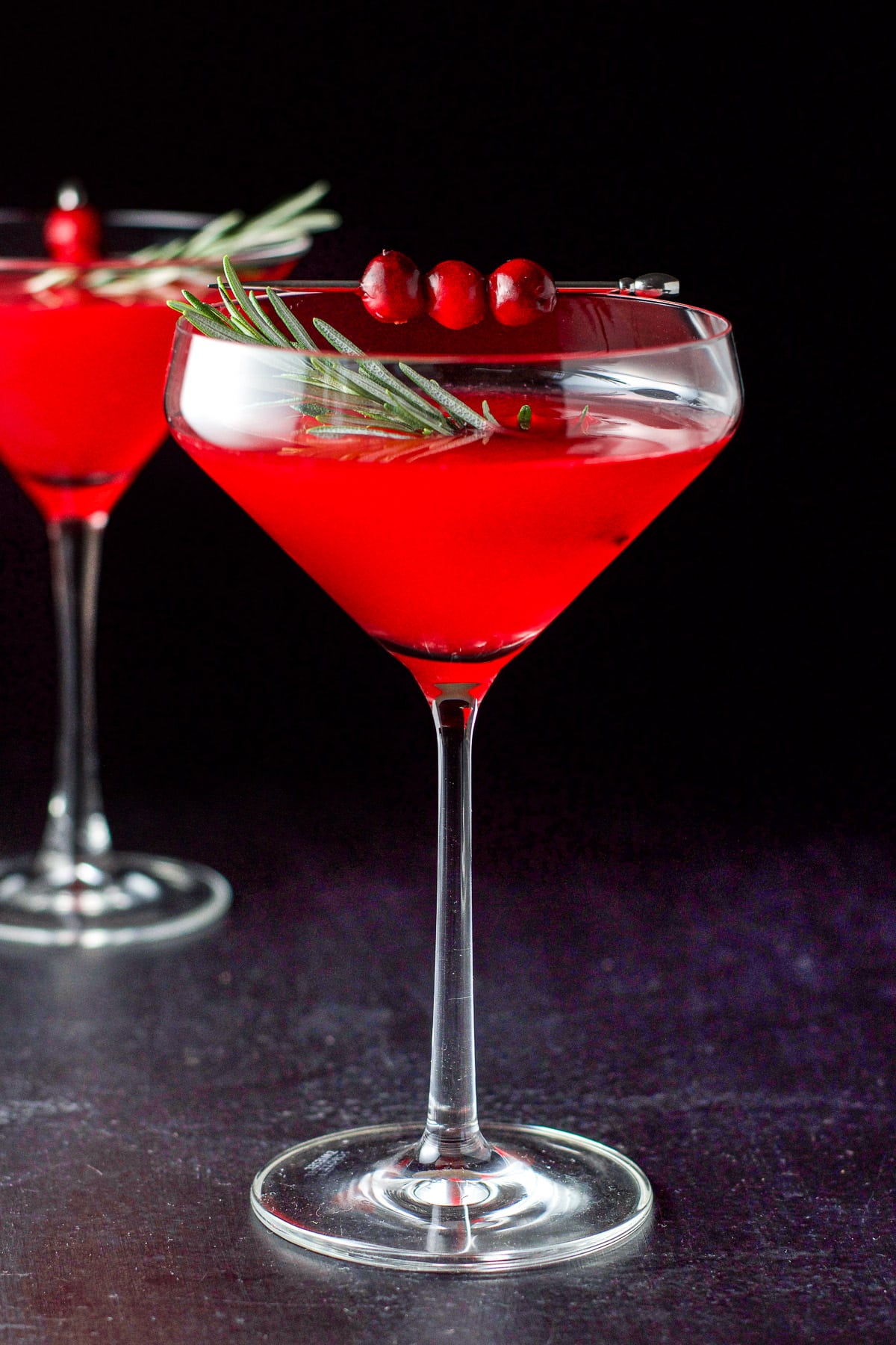 Cranberry cocktail in two martini glasses with the curved glass in front and garnished with cranberries and fresh rosemary