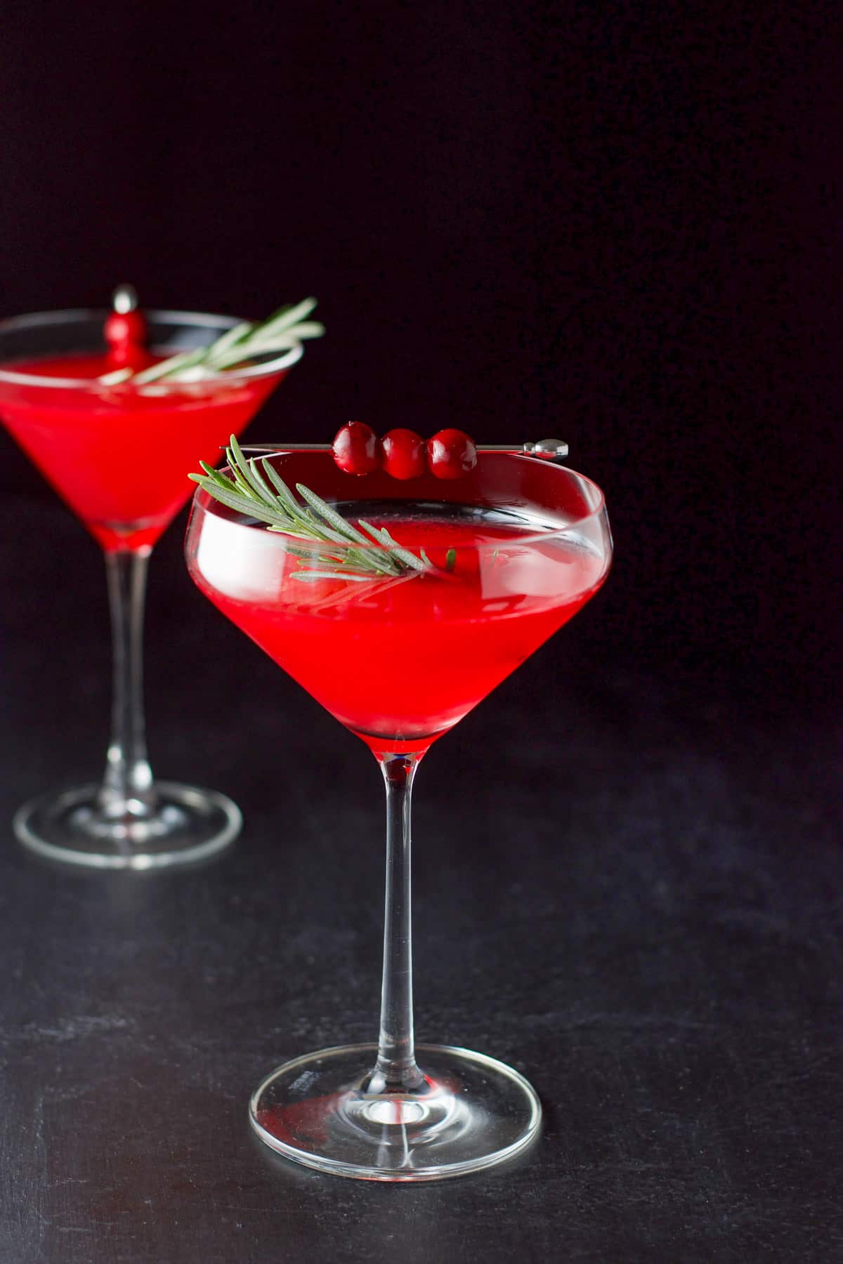 Fun curved martini glass filled with the red cosmo with fresh rosemary as a garnish along with some cranberries on a pick