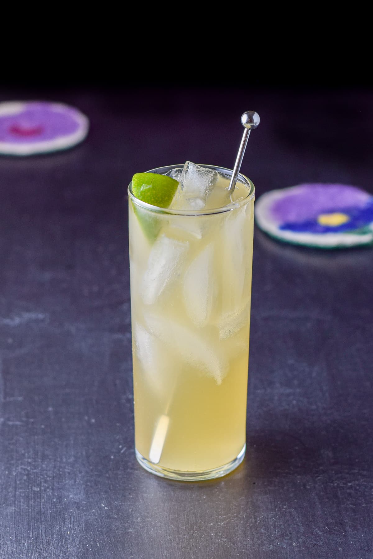 A collins glass filled with the cocktail with a stirrer and lime wedge in it