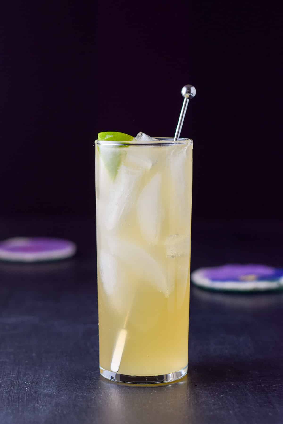 Vertical view of a collin glass with the yellow cocktail with a lime wedge and stirrer in it