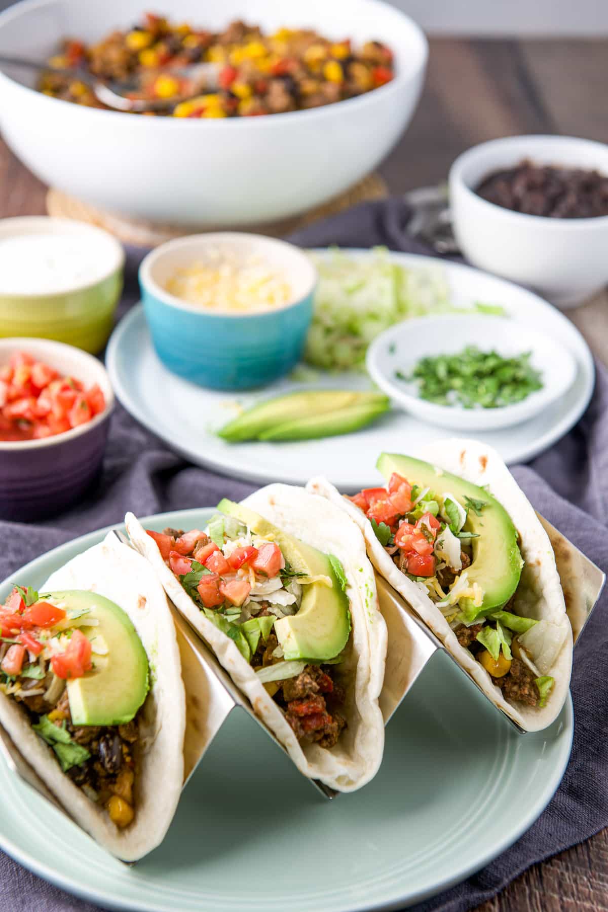 Three soft tacos in a taco holder with ground beef and all the fixings both in the taco and in the back