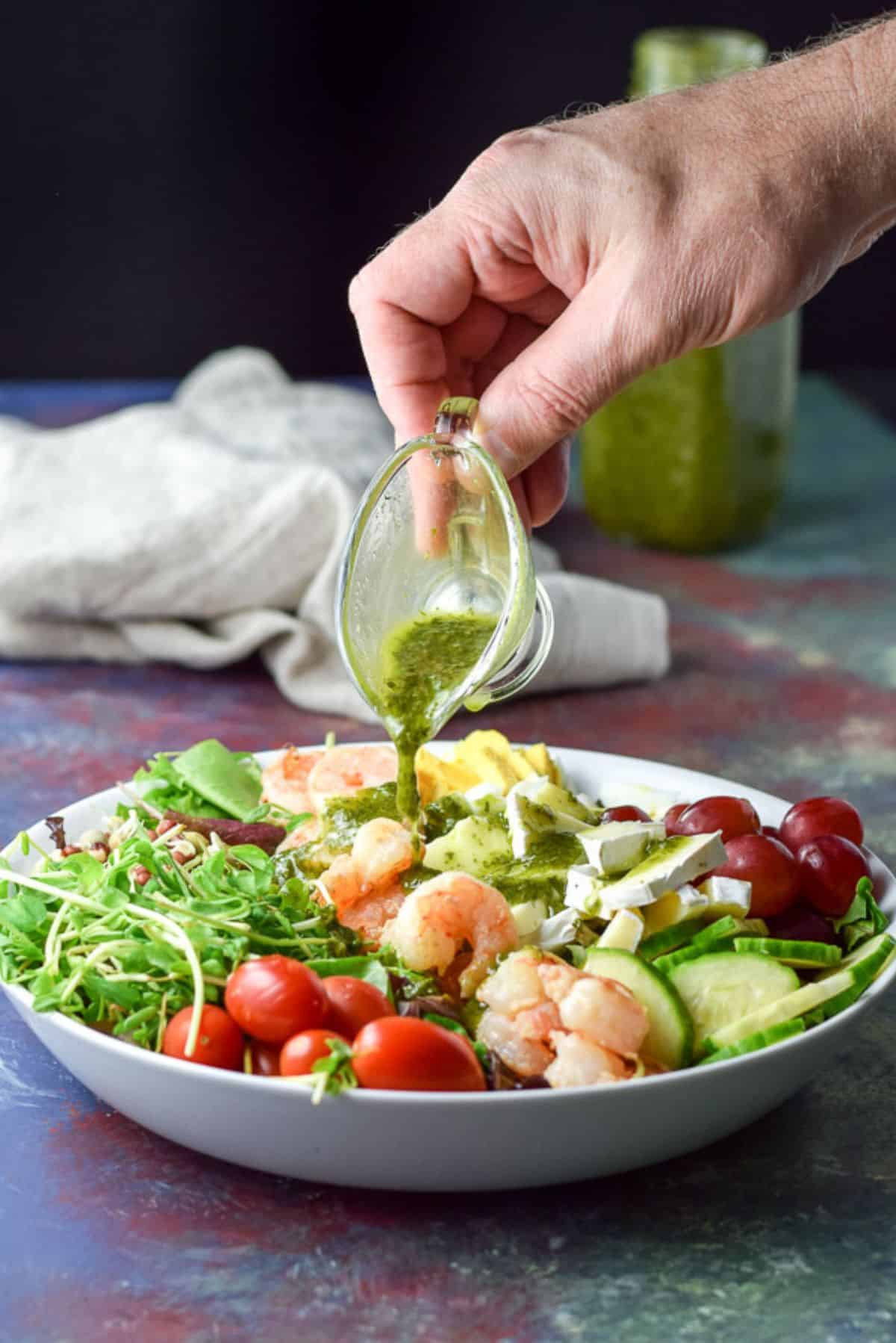 A hand with a container of chimichurri dressing being poured on the salad