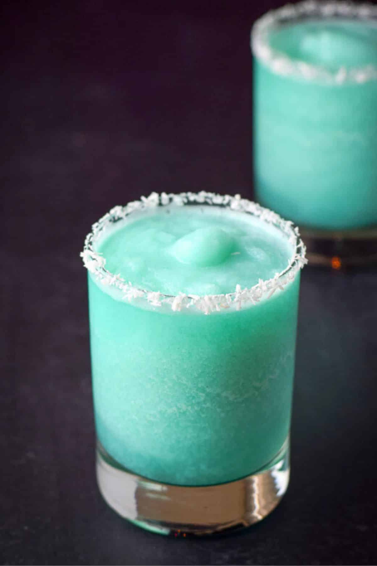 Close up of the blue cocktail in two coconut rimmed glasses on a black table with some lights in the background