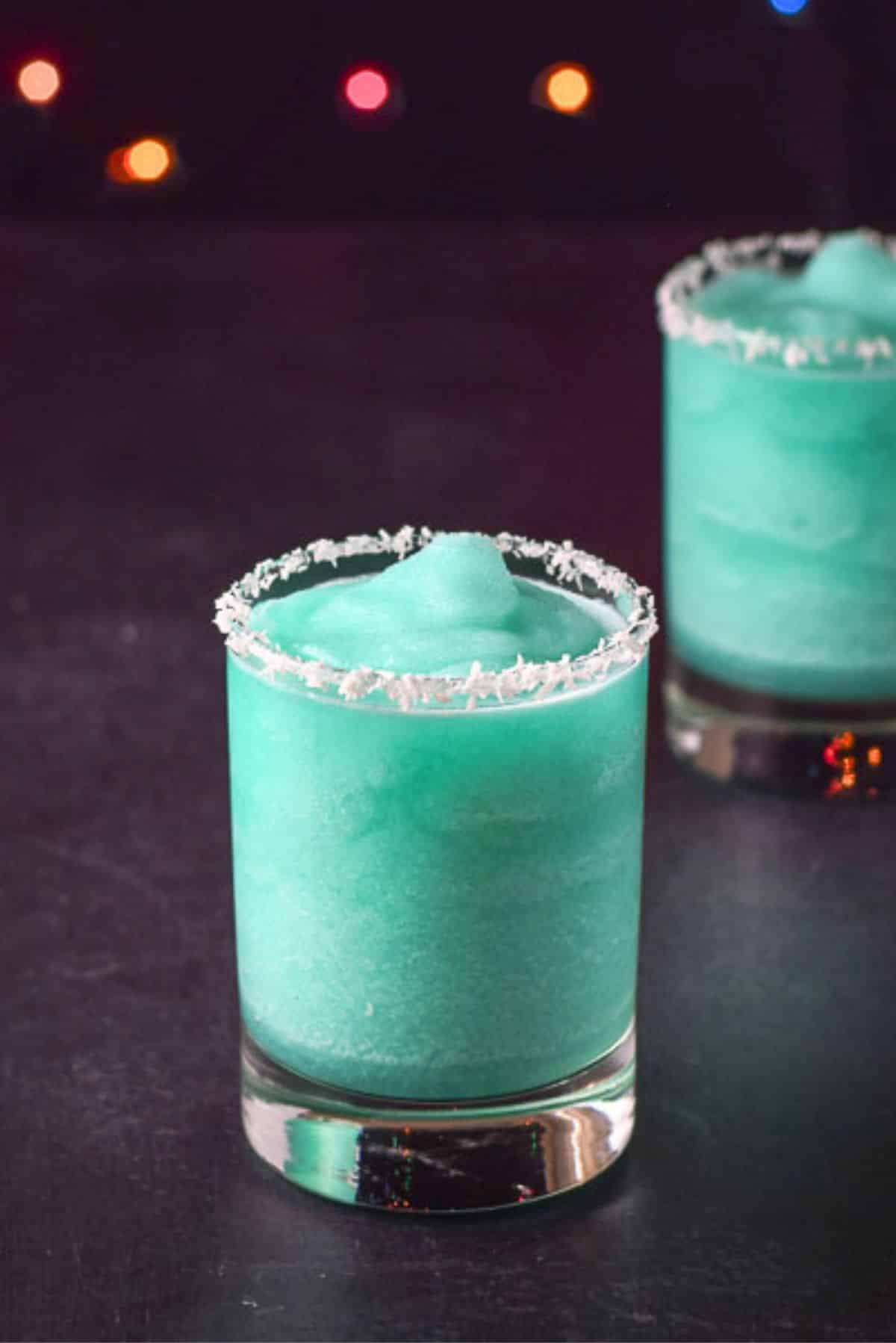 Two Jack Frost cocktails with coconut rims on a black table and colorful lights behind them