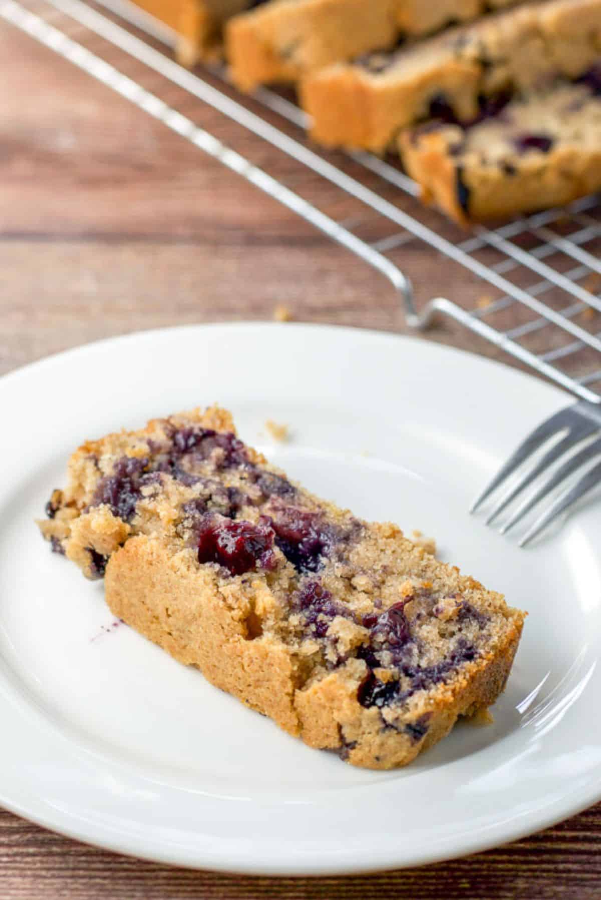 A piece of lemon blueberry bread on a plate with a few pieces on a wire rack behind it