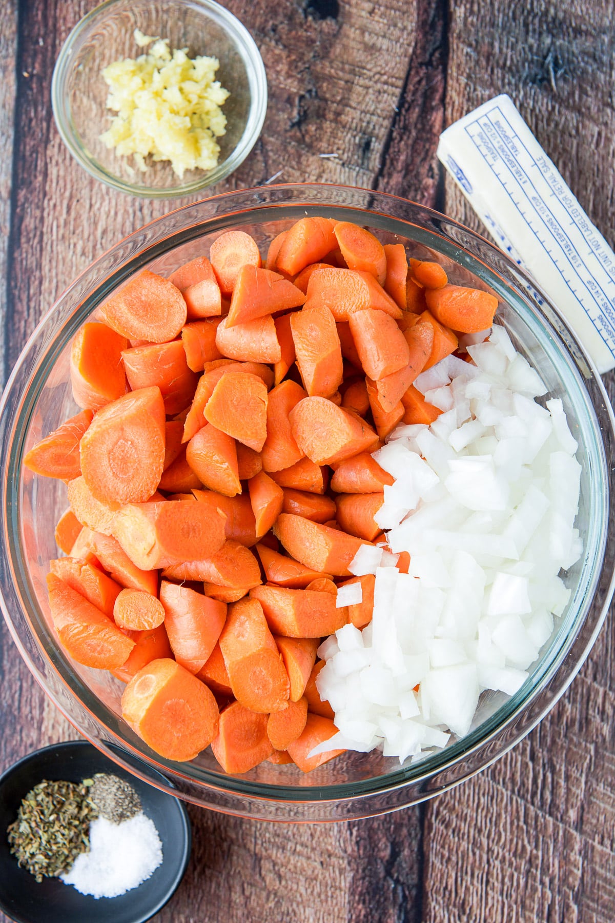 Overhead view of cut carrots and onion in a glass bowl, butter, crushed garlic, basil, pepper, and salt
