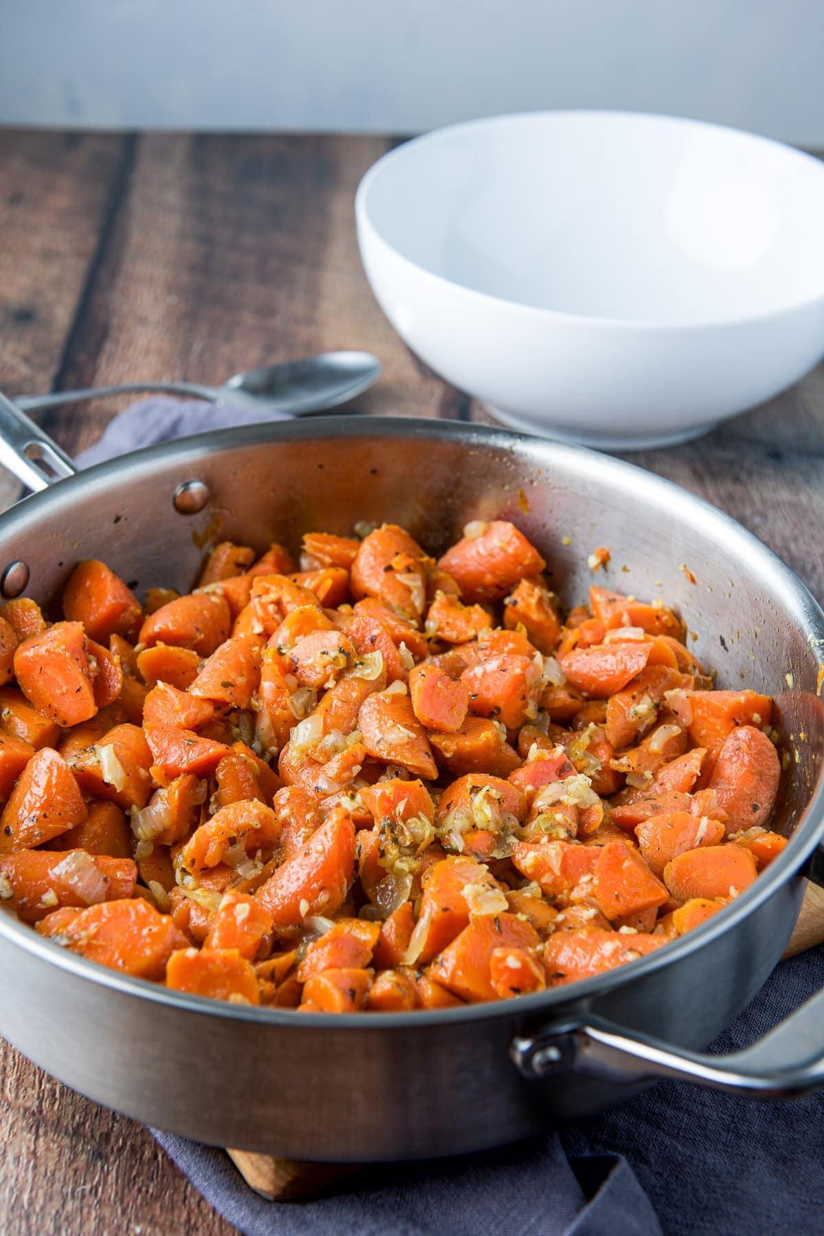 A metal saute pan with the cooked carrots in it in front of a spoon and a serving bowl