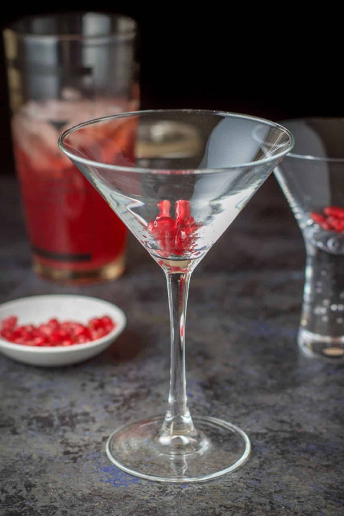 Pomegranate seeds spooned into the martini glasses with more seeds in the background along with the cocktail shaker