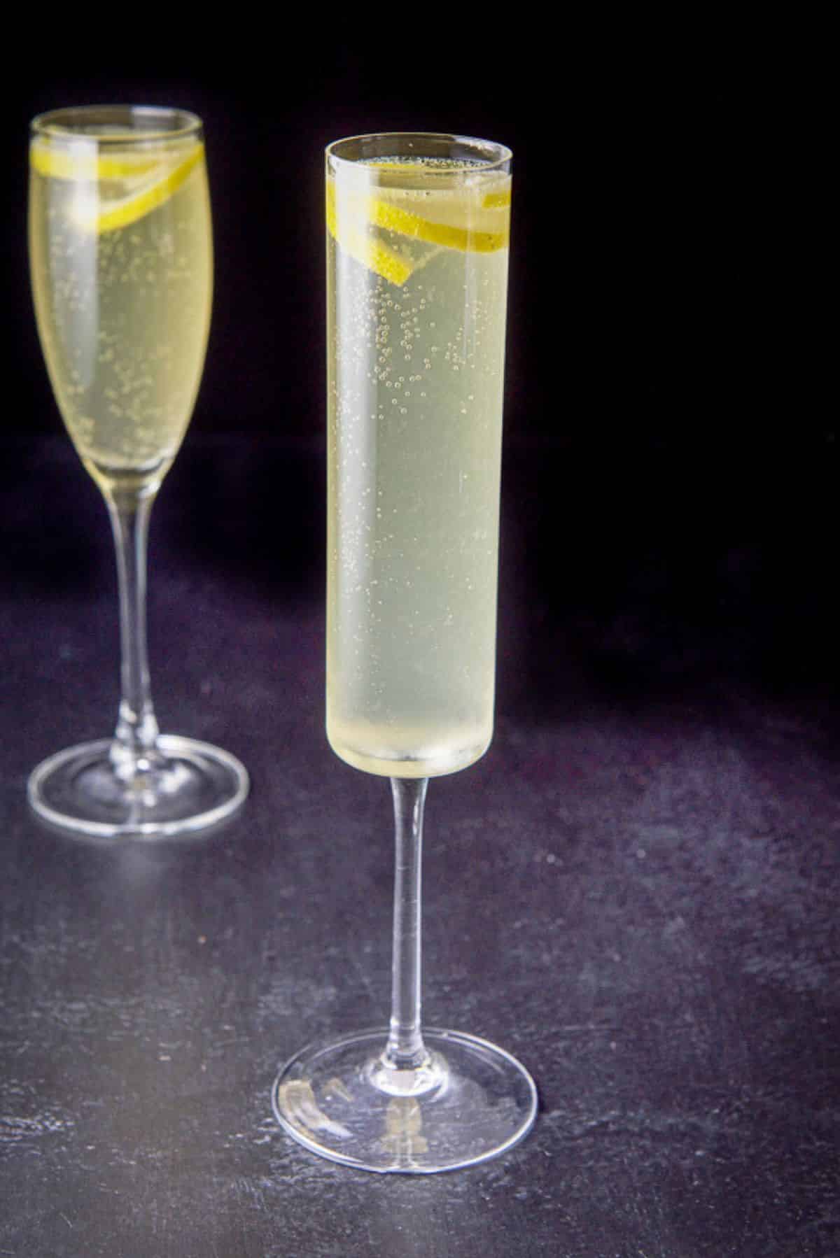 The thin champagne glass filled with the drink, the classic glass is behind it and both have lemon twists in the glasses