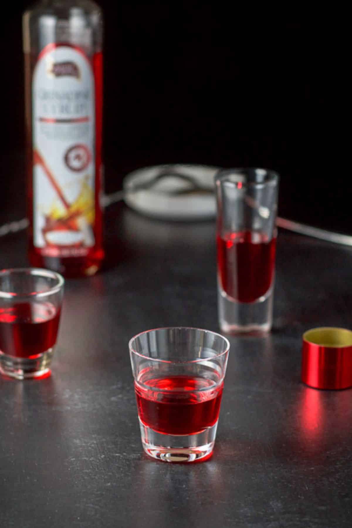 Grenadine poured into the three shot glasses with the bottle and spoons in the background