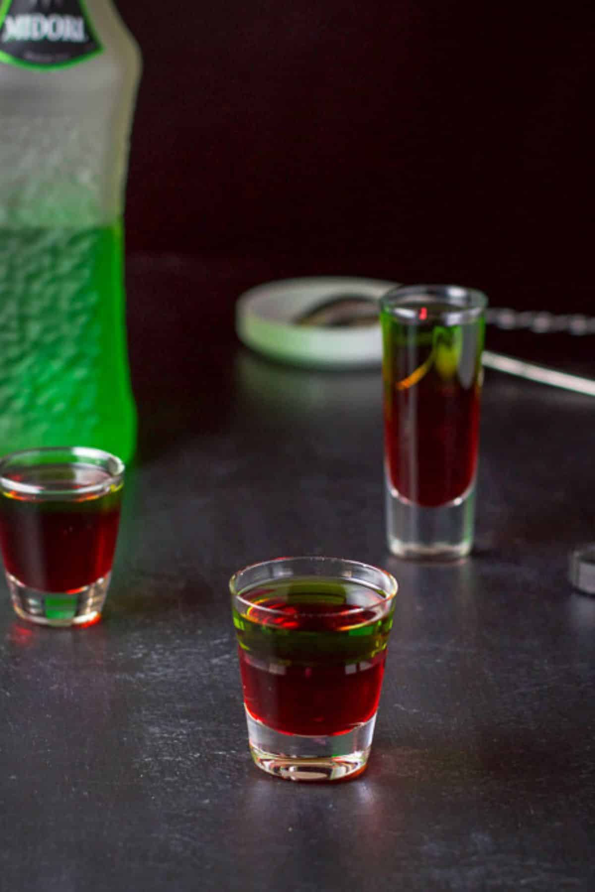 The Midori layered into the shot glasses with the bottle and spoons in the background