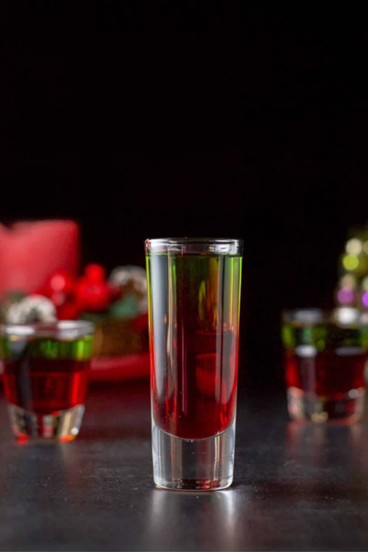 Vertical view of the tall glass filled with the green and red shot. There is a Christmas tree and candle in the background