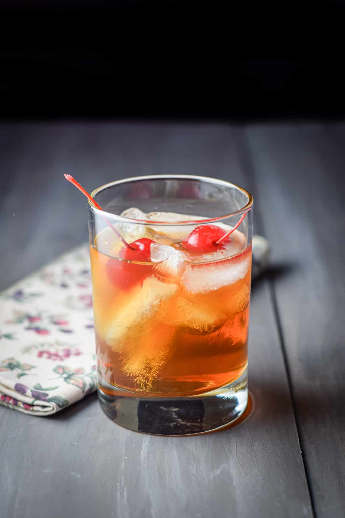 A grey cocktail with a glass with the whiskey manhattan with two cherries