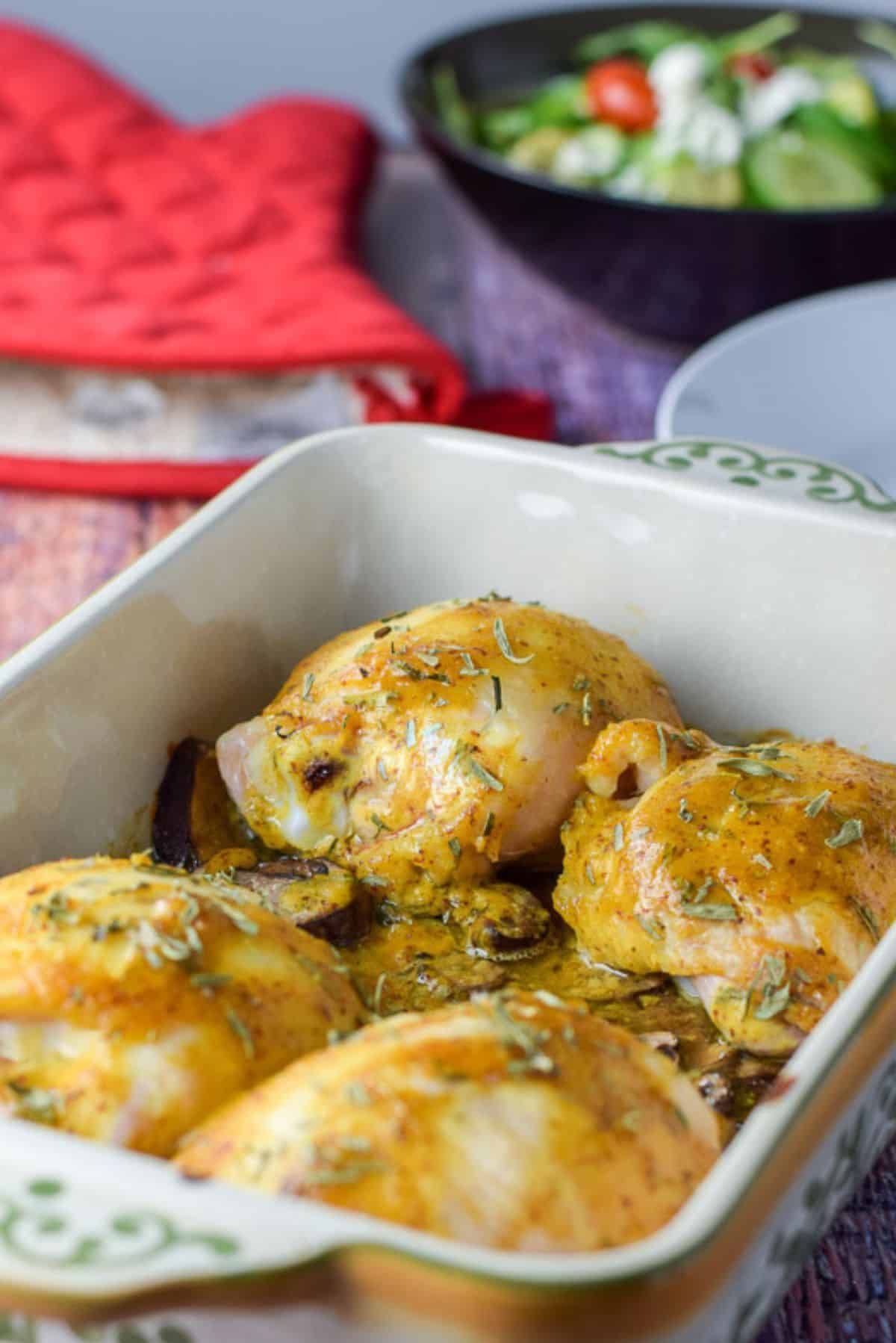 A baking dish with the mustard thighs straight out of the oven with a salad and oven mitt in the background