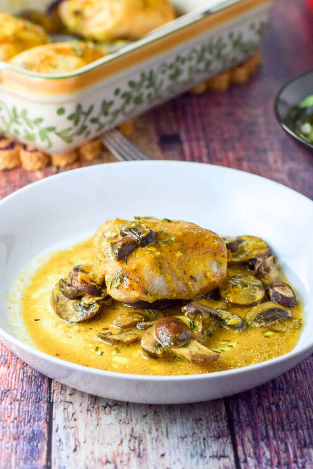 A white shallow bowl filled with a chicken thigh with sauce and mushrooms in it. There is a baking dish in the background
