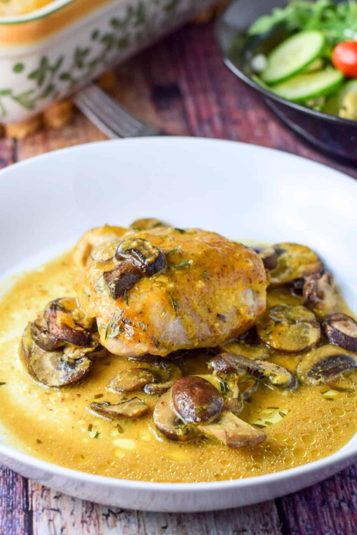 Closer look of a shallow bowl filled with saucy chicken and mushrooms. There is a baking dish and a salad in the background