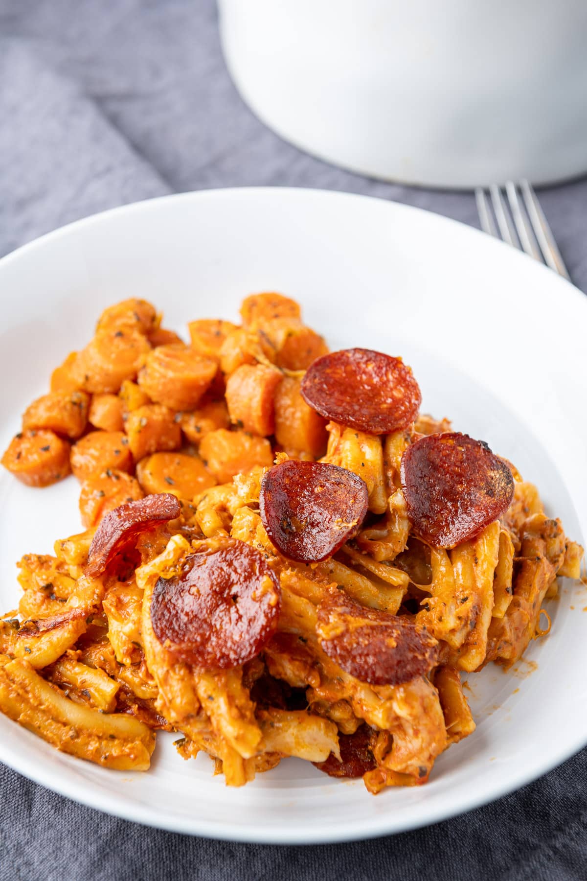 A white plate of pasta, pepperoni and cooked carrots