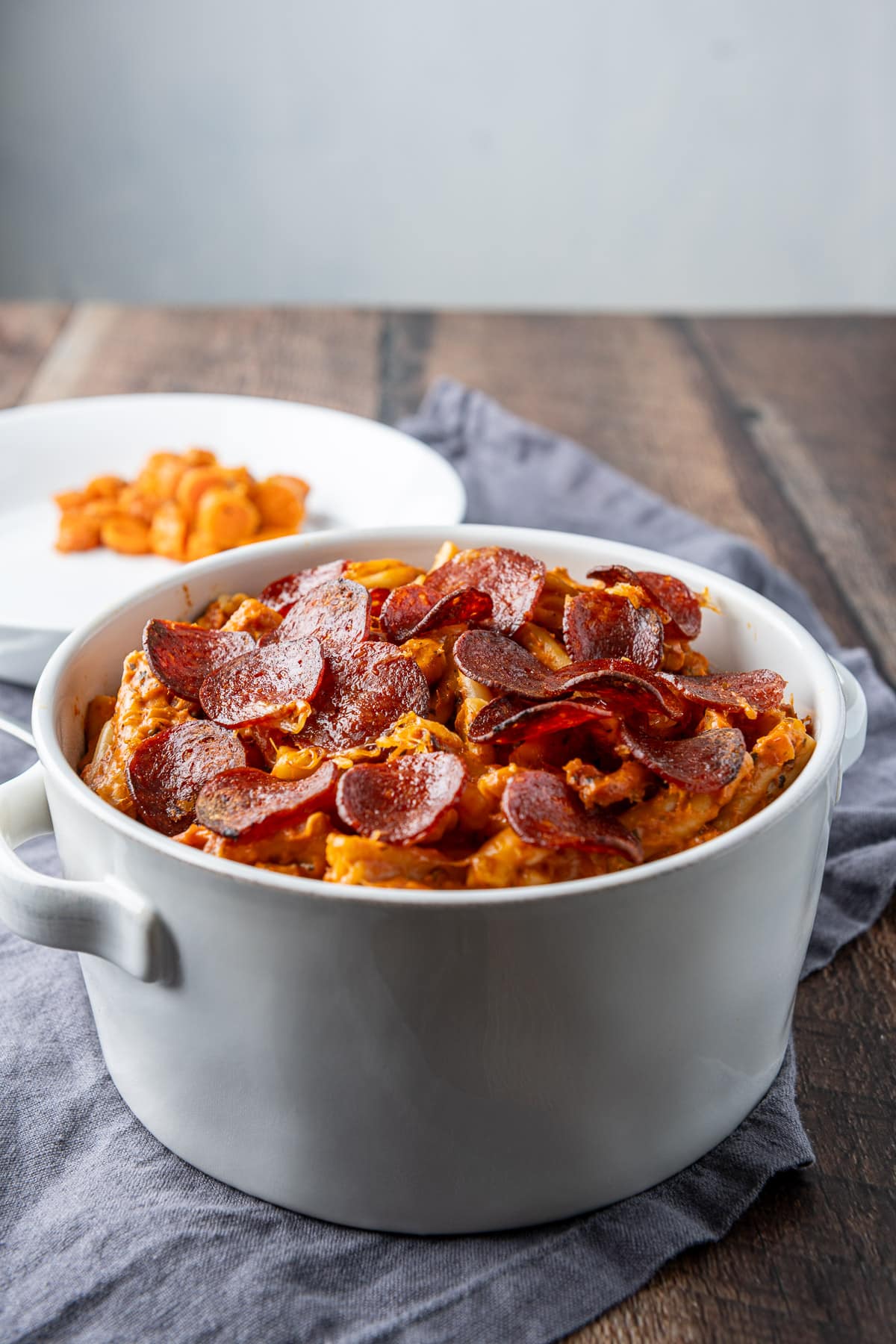 A white pan with the pasta and pepperoni in it with a plate with some carrots in back