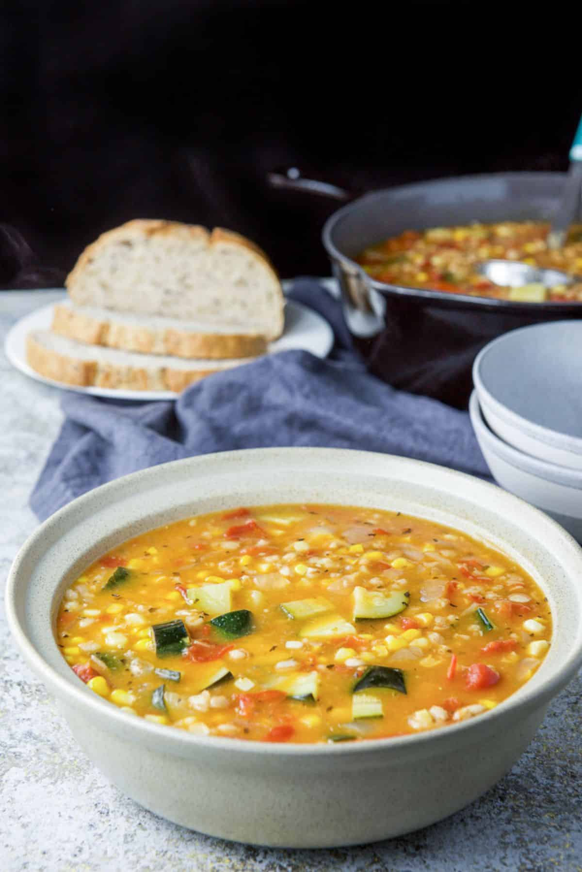 Soup with corn and zucchini in a serving bowl with the bread, a pan and bowls in the background