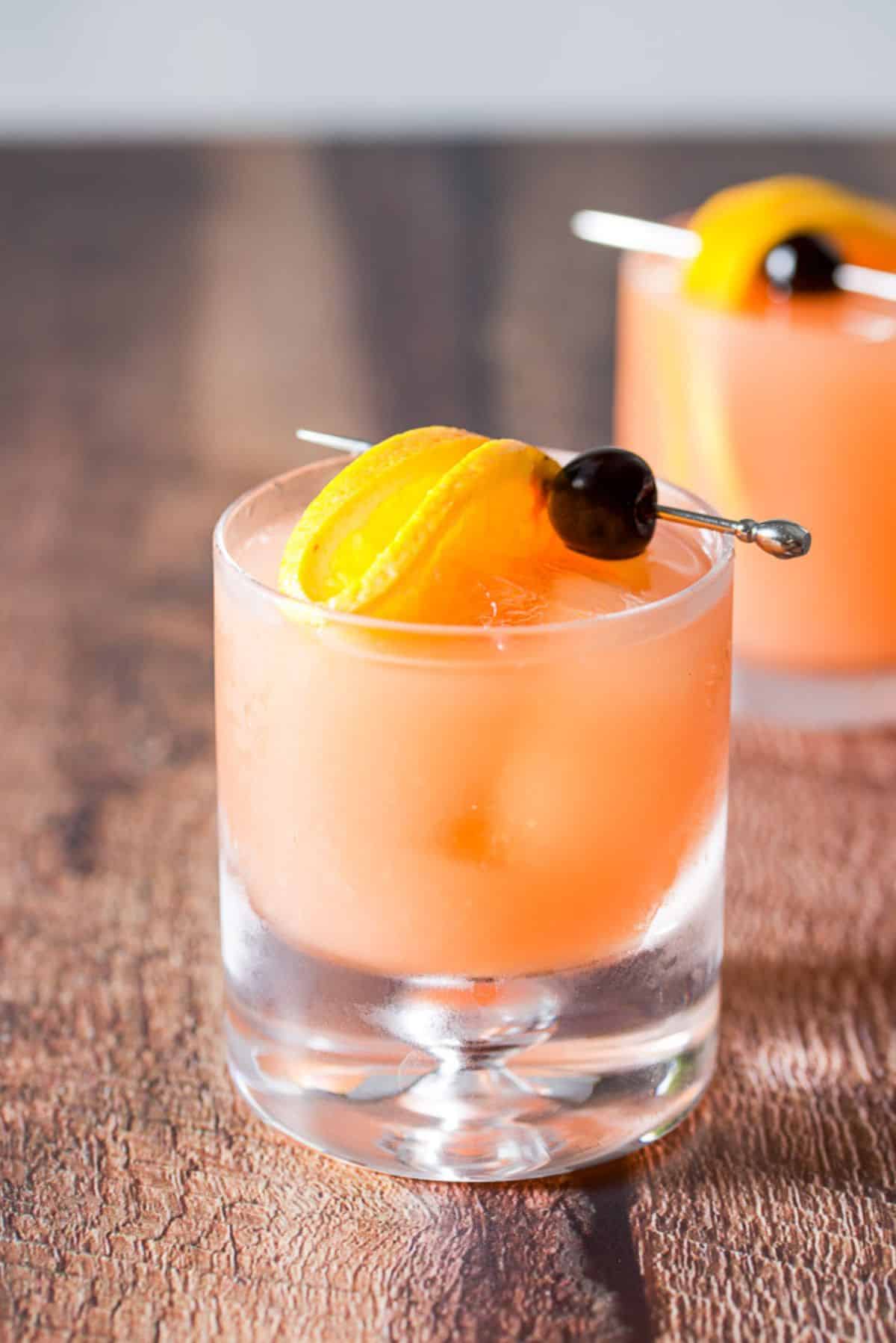 Two glasses on a wooden table with the mai tai in it along with cherries and orange slices