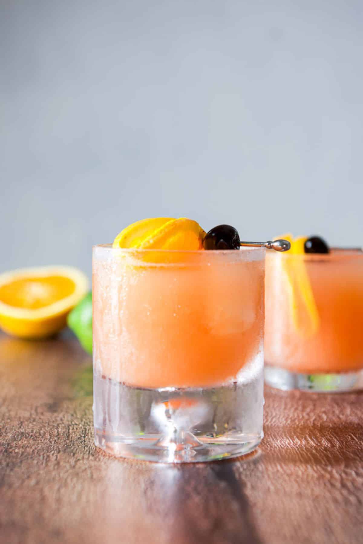 Vertical view of two glasses with the orange drink along with garnish and an orange on the table