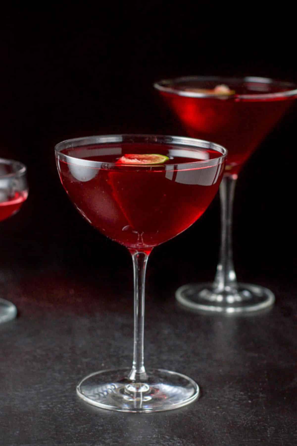 The red cocktail poured into 3 glasses - a bowl glass, classic martini and coupe. There are limes floating in the glasses
