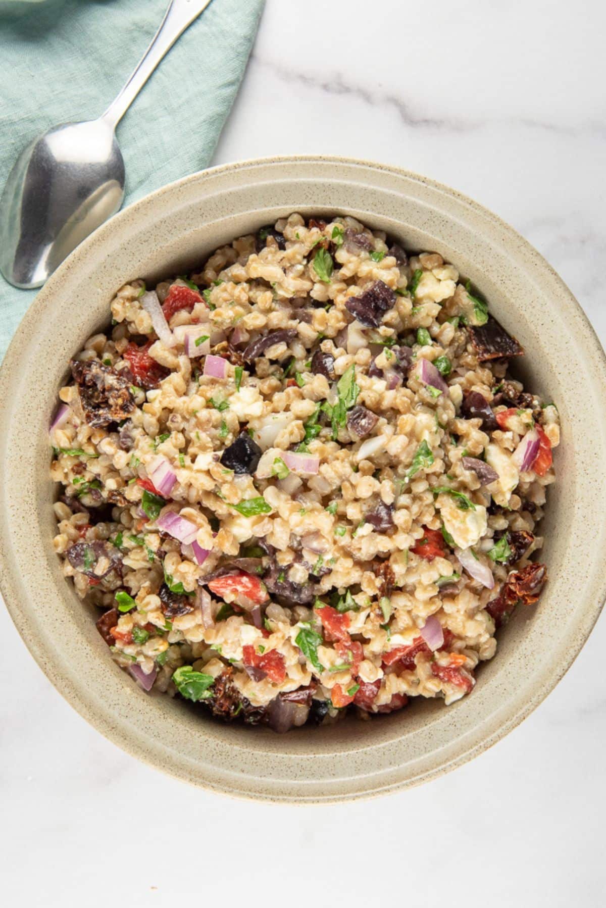 Overhead view of the grain salad in a tan bowl. There is a spoon and green napkin also