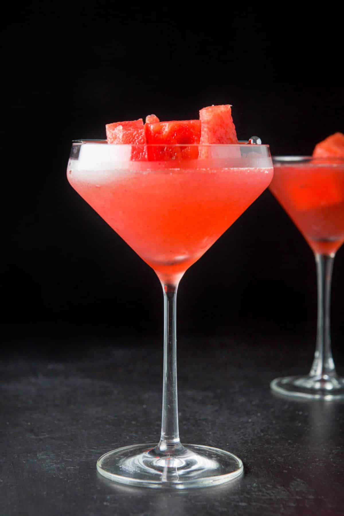 vertical view of a unique martini glass with a cosmo in it along with watermelon chunks