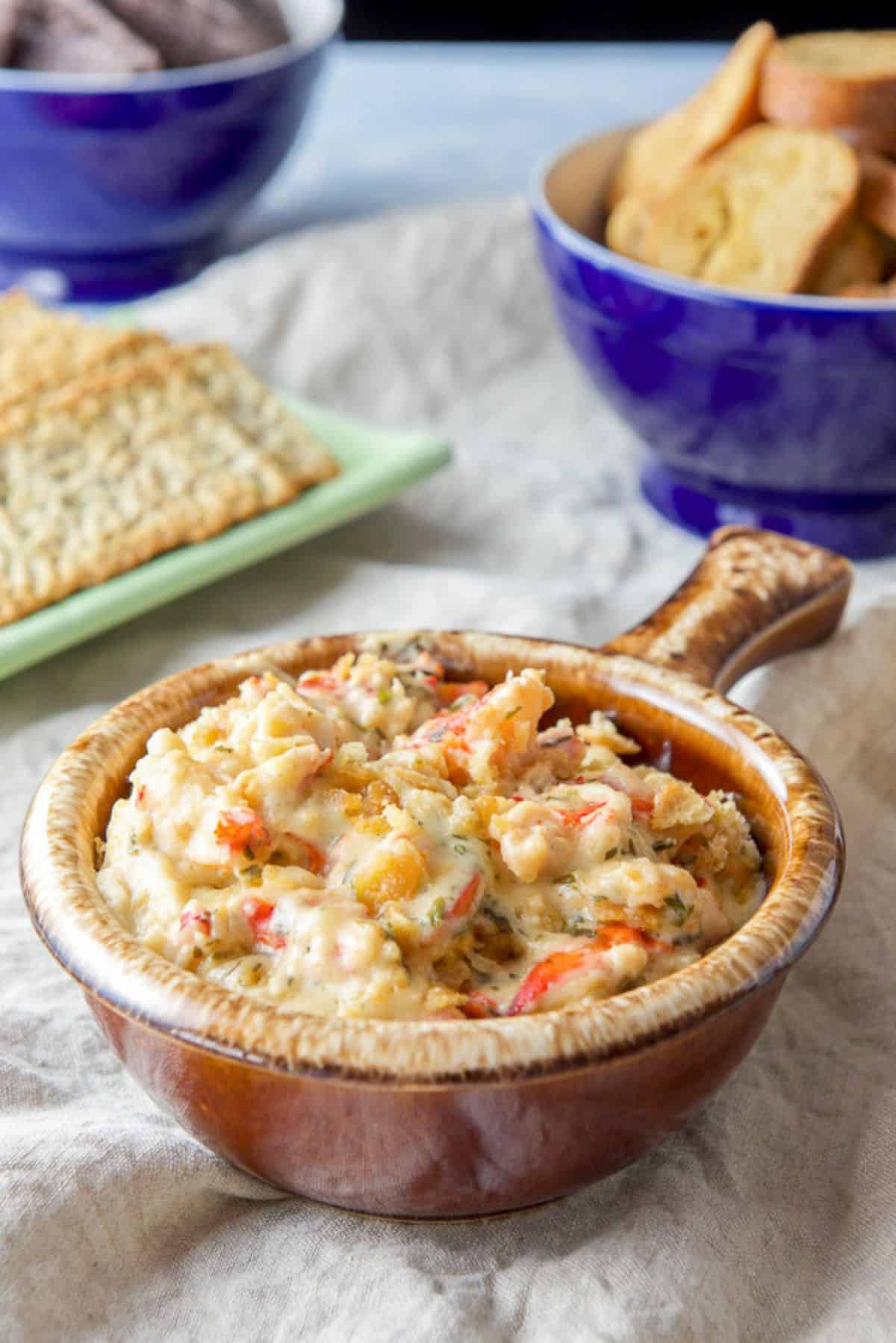 A brown crock filled with a dip with lobster poking out. There are crackers, chips, and bread crisps in the background