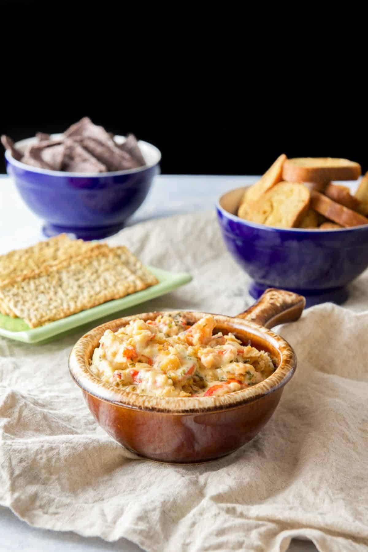A brown crock filled with a chunky lobster dip with crackers, bread crisps and chips in the background