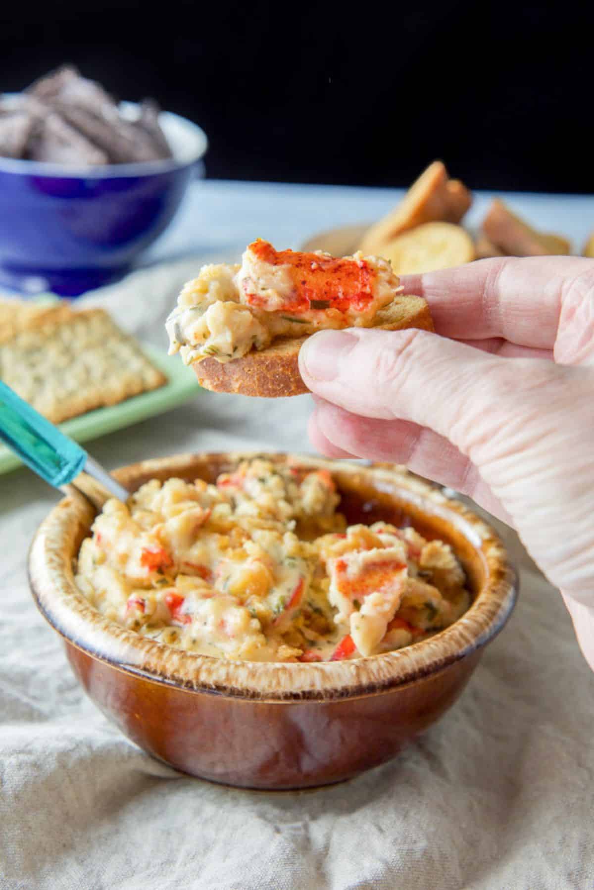 A hand holding bread with a lobster mix on it over a bowl of the casserole