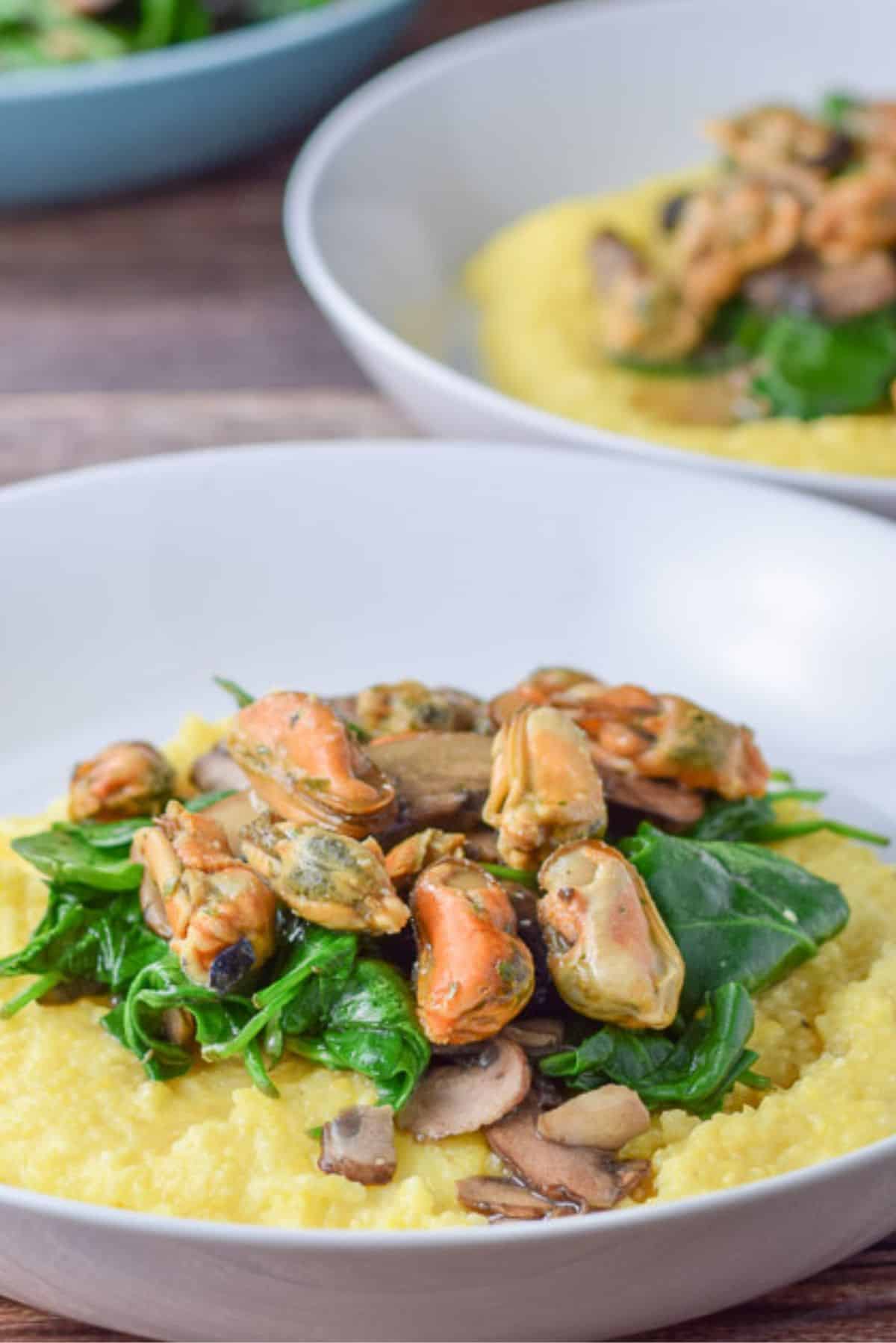 Close up of a white deep dish with polenta, veggies and garlic mussels on top