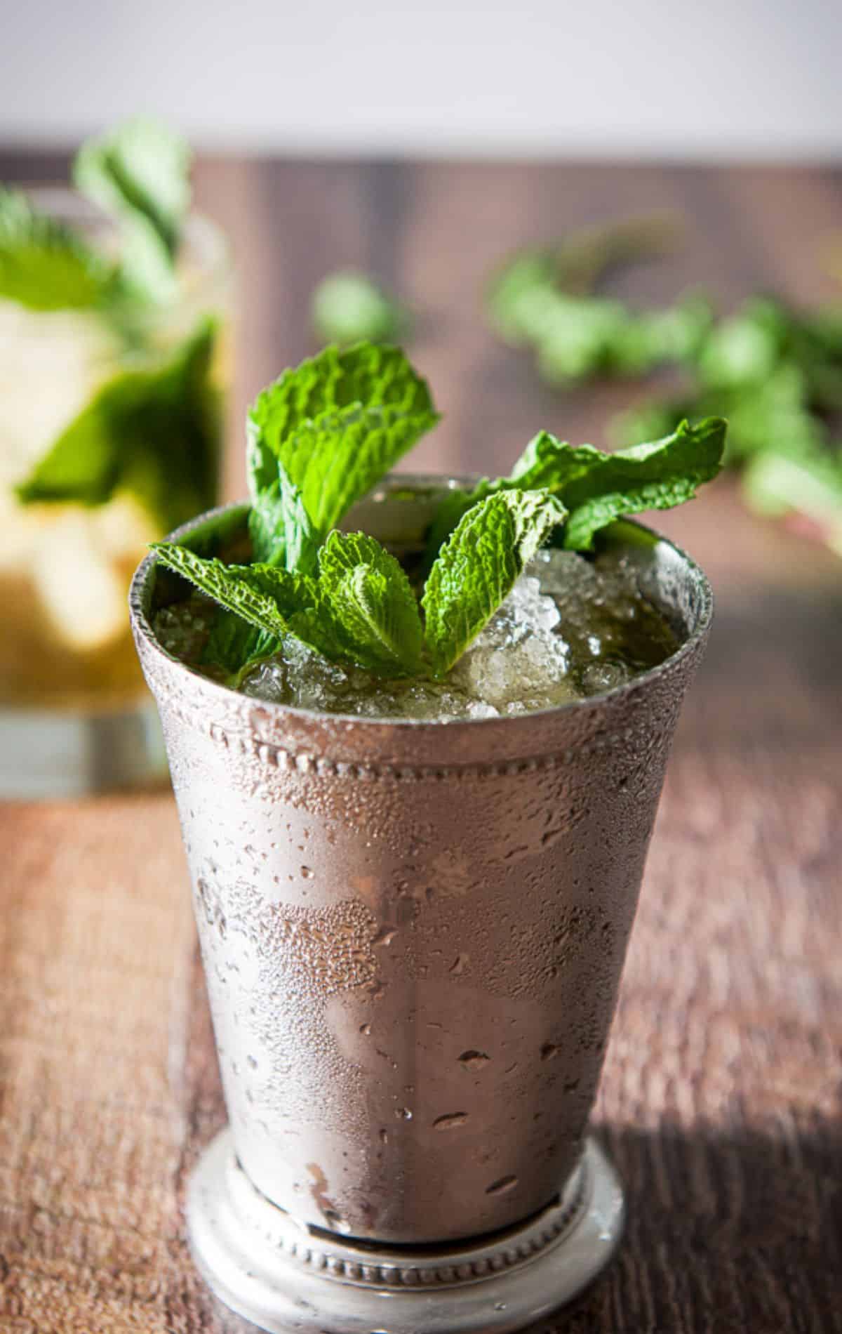 A metal cup in front of a glass filled with the mint julep with lots of fresh mint as garnish