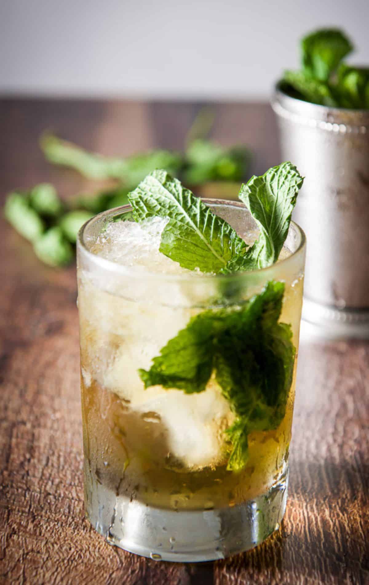 A glass filled with ice and the mint cocktail with a metal glass in the back and fresh mint on the table
